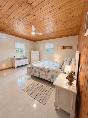 Un dormitorio con techo de madera, una cama con colcha floral, una mesita de noche, un aparador y dos ventanas con persianas.