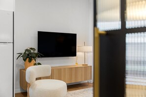 A modern living room with a flat-screen TV mounted on the wall, a wooden sideboard, a white upholstered chair, a floor lamp, and a potted plant. - Luna—Canopy Views from a Peaceful Garden Haven (College Park)