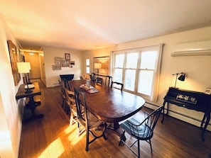 Comedor con una gran mesa de madera rodeada de sillas, un piano y una ventana con cortinas.