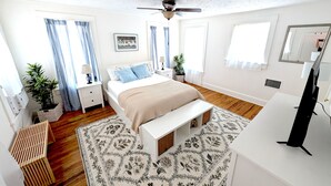 Quarto com cama, mesas de cabeceira, ventilador de teto, televisão e uma grande janela com cortinas.