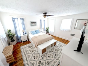 Un dormitorio con cama, mesitas de noche, ventilador de techo, televisión y un gran ventanal con cortinas.