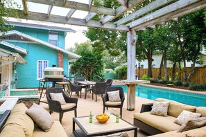 Un área de patio techada con mobiliario exterior, una piscina y una casa de fachada azul.