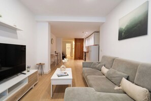 Ein modernes Wohnzimmer mit einem Flachbildfernseher, einem grauen Sofa, einem Couchtisch mit Büchern und einem Essbereich im Hintergrund.