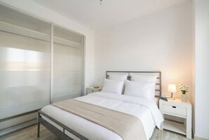 Une chambre à coucher moderne avec une grande porte coulissante, un lit recouvert de linge de maison blanc, une table de chevet avec une lampe et une commode avec des livres et un vase de fleurs.