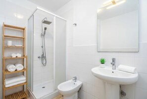 Une salle de bain moderne dotée d’une douche à parois de verre, d’un lavabo sur colonne, d’un bidet et d’une étagère en bois où sont posées des serviettes.