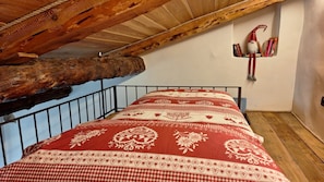 A bedroom with a sloped ceiling, a metal bed frame, a patterned bedspread, and a small shelf with books and a gnome decoration. - I Sitoù - Ratizin Apartment: The Scent of Ancient Wood (Ostana)