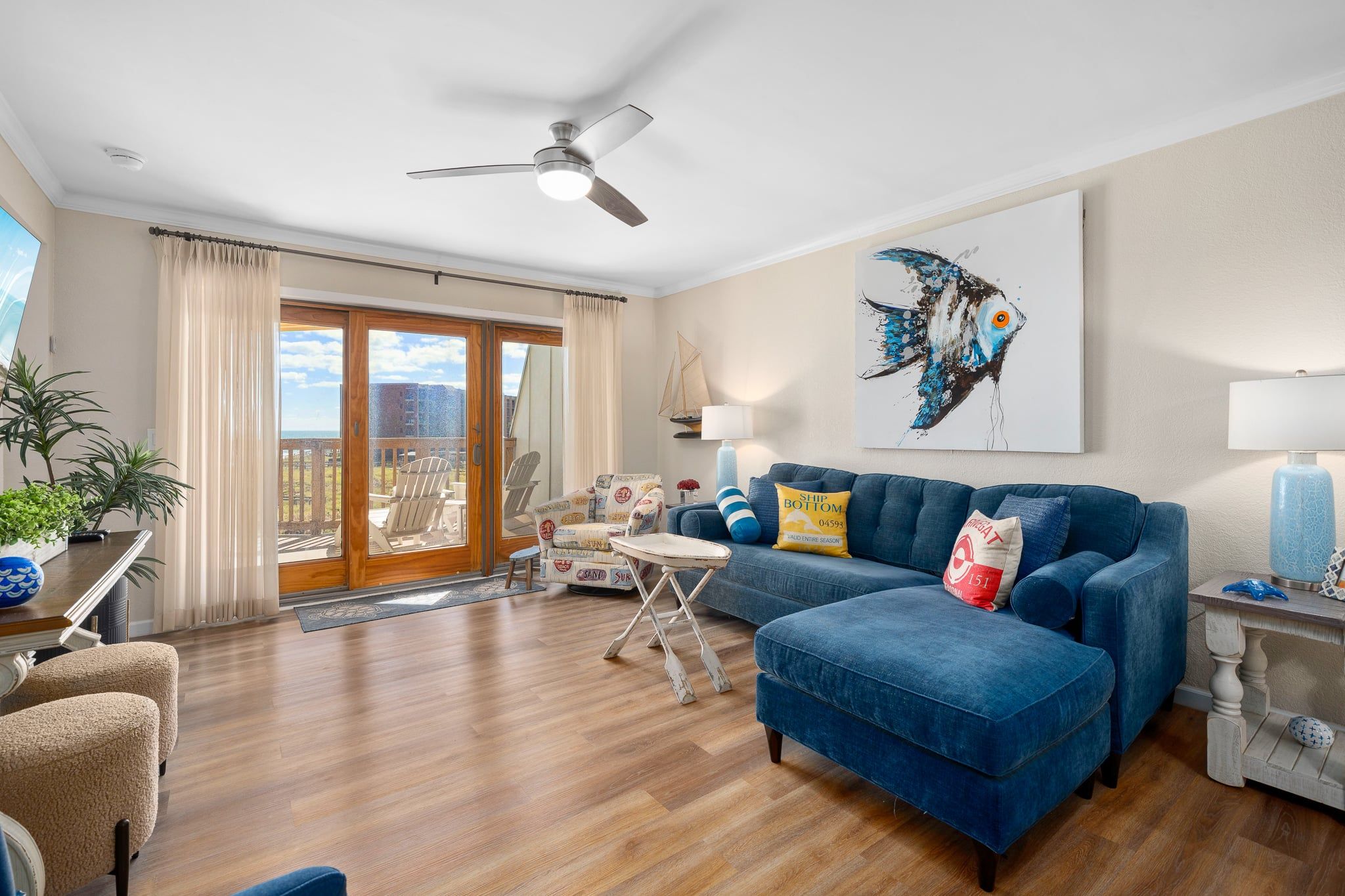 Un salon avec un canapé bleu, un ventilateur de plafond, un tableau représentant un poisson et une vue sur l’extérieur, visible à travers une porte vitrée.