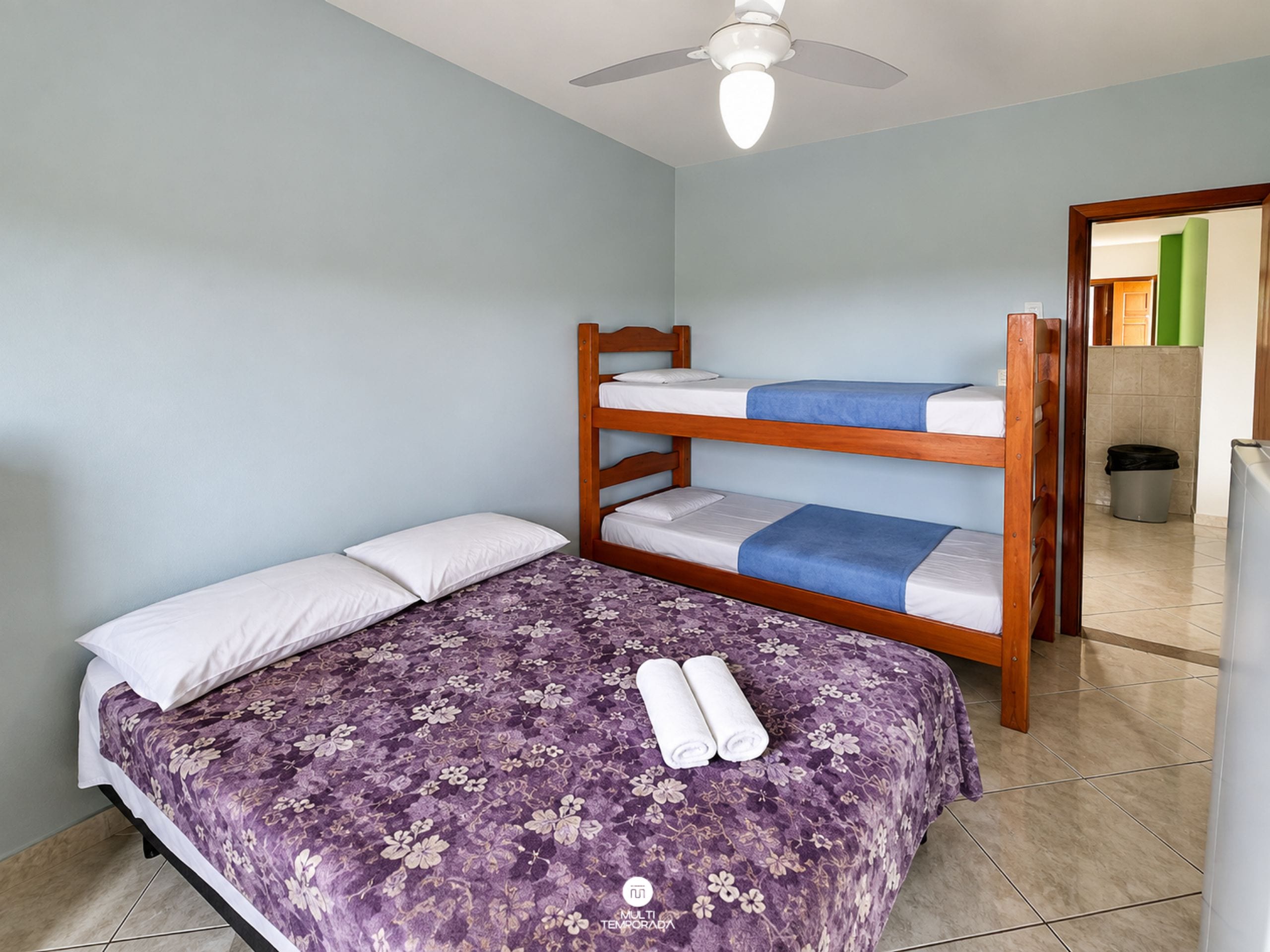 Una cama individual con una colcha floral y dos almohadas, una cama litera con una manta azul, y un ventilador de techo en una habitación con paredes azul claro y suelo de baldosas.