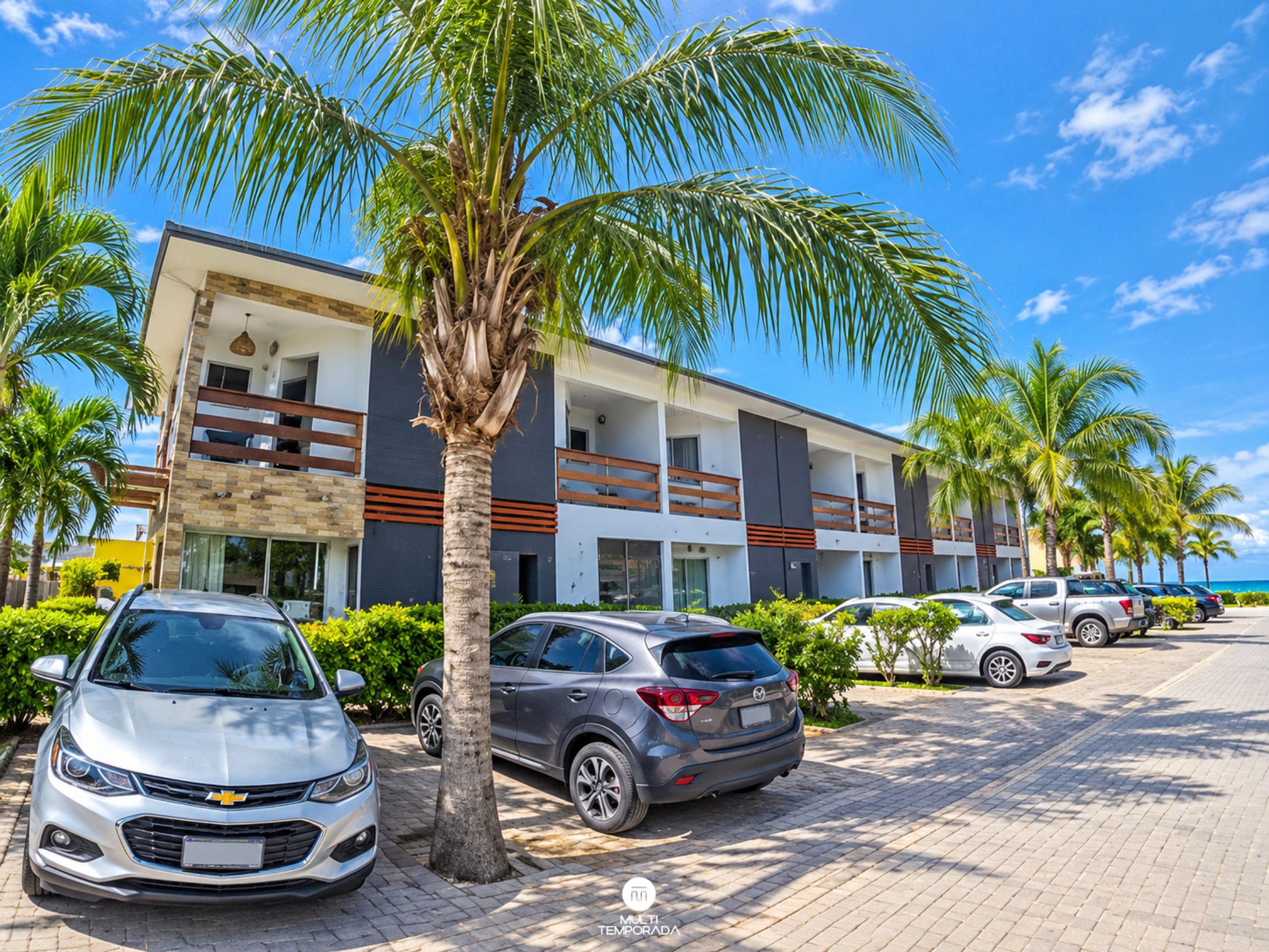 A hotel with a modern design, featuring a two-story structure with balconies, parked cars, and palm trees.