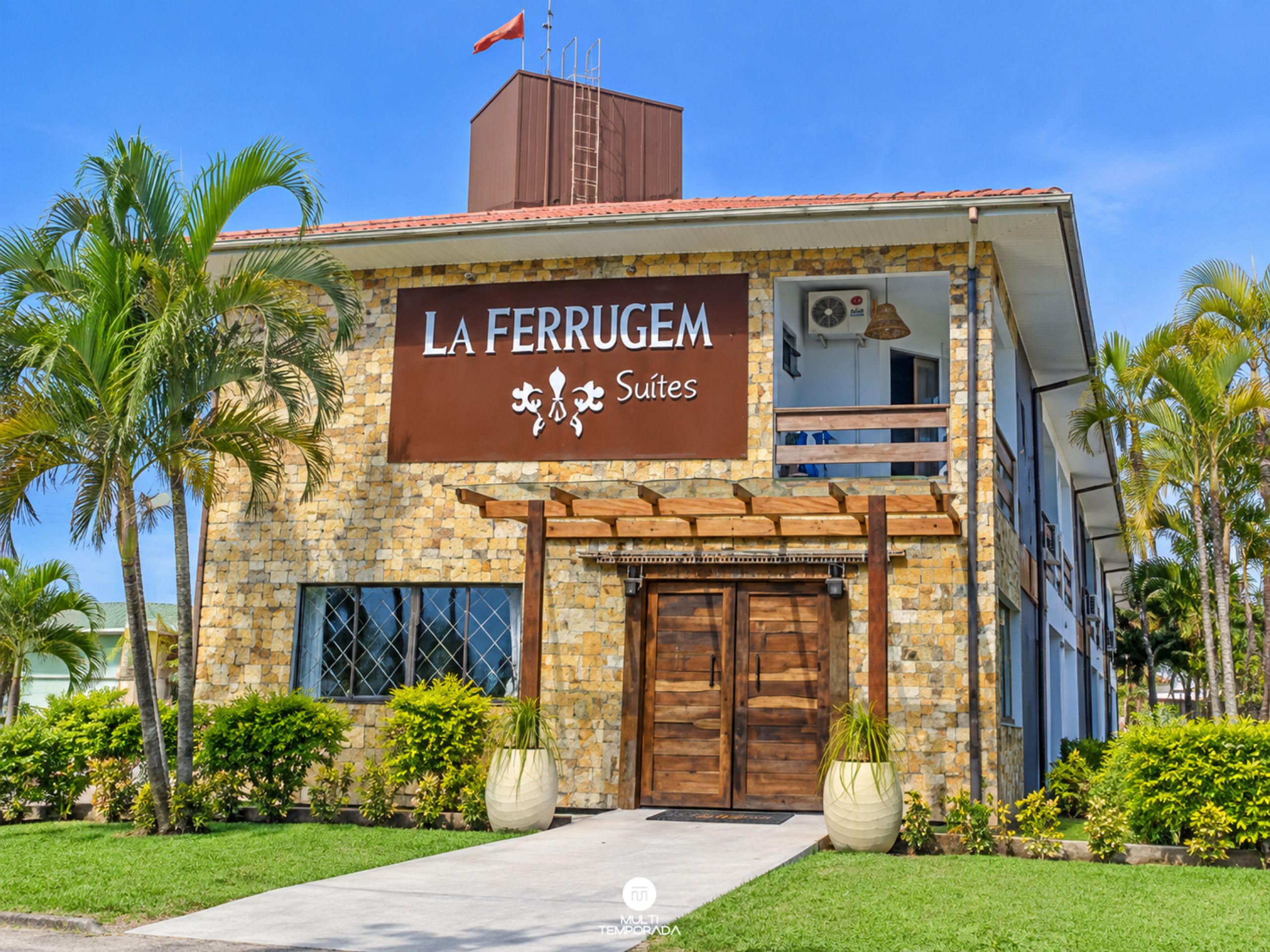 Un hotel llamado 'La Ferrugem Suites' con fachada de piedra, ventanas grandes y un balcón.