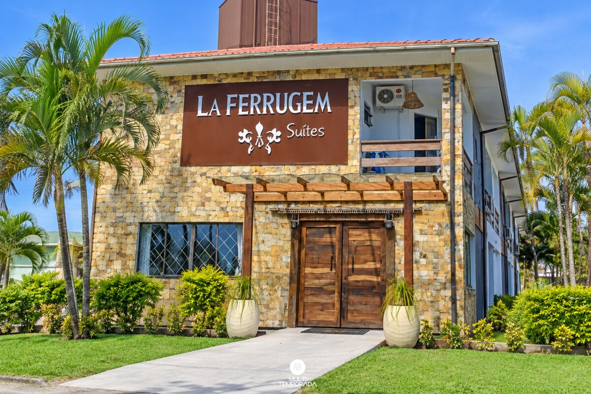 Um hotel chamado 'La Ferrugem Suites' com fachada de pedra, janelas amplas e uma varanda.