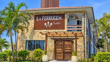 Un hotel llamado 'La Ferrugem Suites' con fachada de piedra, ventanas grandes y un balcón.