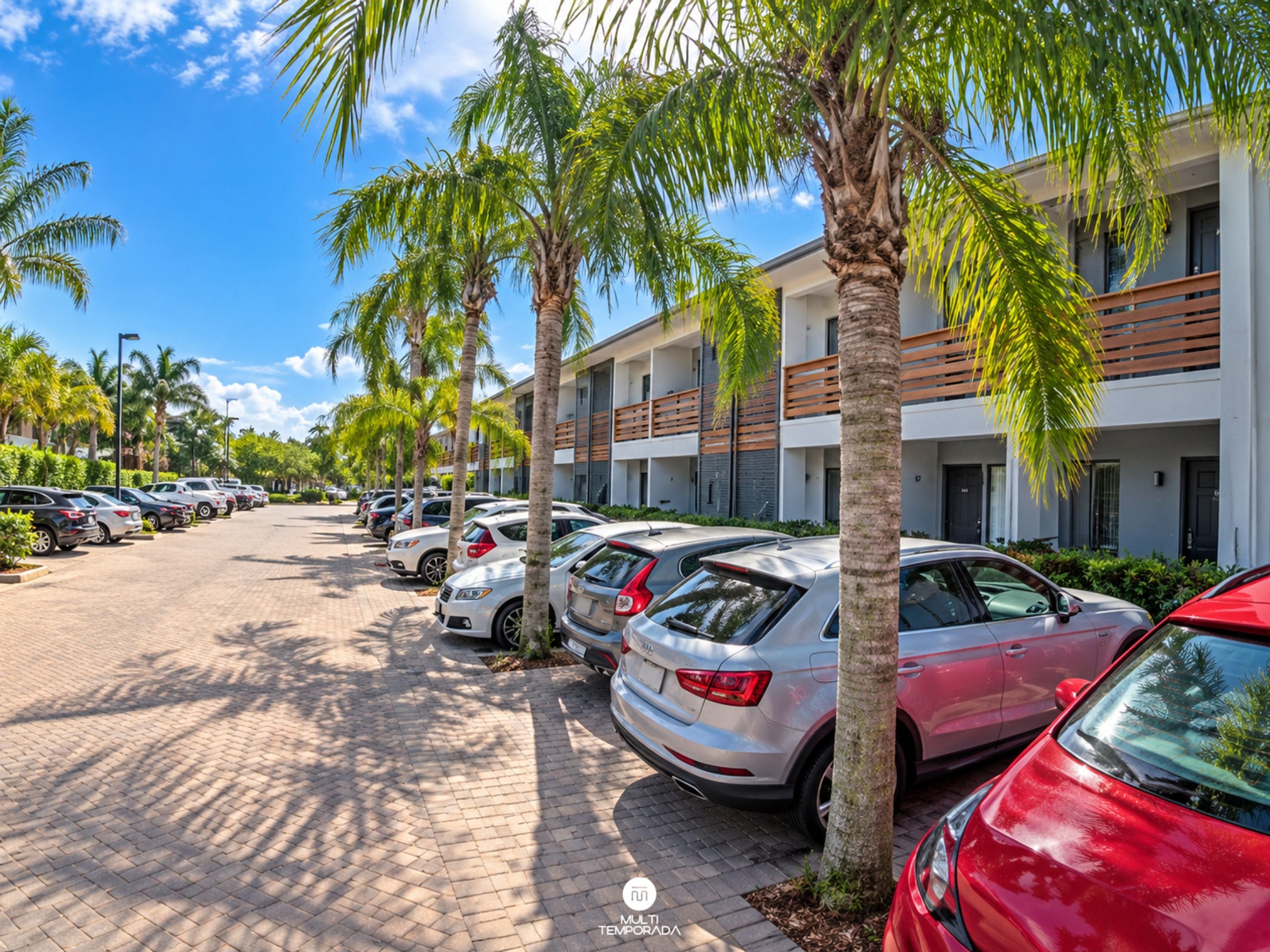 Uma área de estacionamento com vários carros, palmeiras e um edifício com varandas.