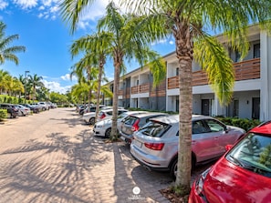 A parking area with multiple cars, palm trees, and a building with balconies. - La Ferrugem Suites - 100m da Praia - Ferrugem/sc (Garopaba)
