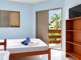 Quarto com cama, televisão, guarda-roupa e porta de vidro deslizante para uma varanda com vista para palmeiras.