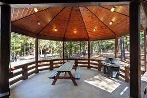 A covered picnic area with a wooden table and grill, surrounded by a wooden fence and trees. - Castaway Lodge - spacious cabin with private rooms (Broken Bow)