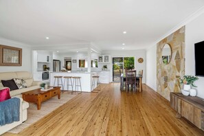 A spacious living area with a kitchen and dining space, wooden flooring, and a stone accent wall. - Aura by Coast Hosting (Terrigal)
