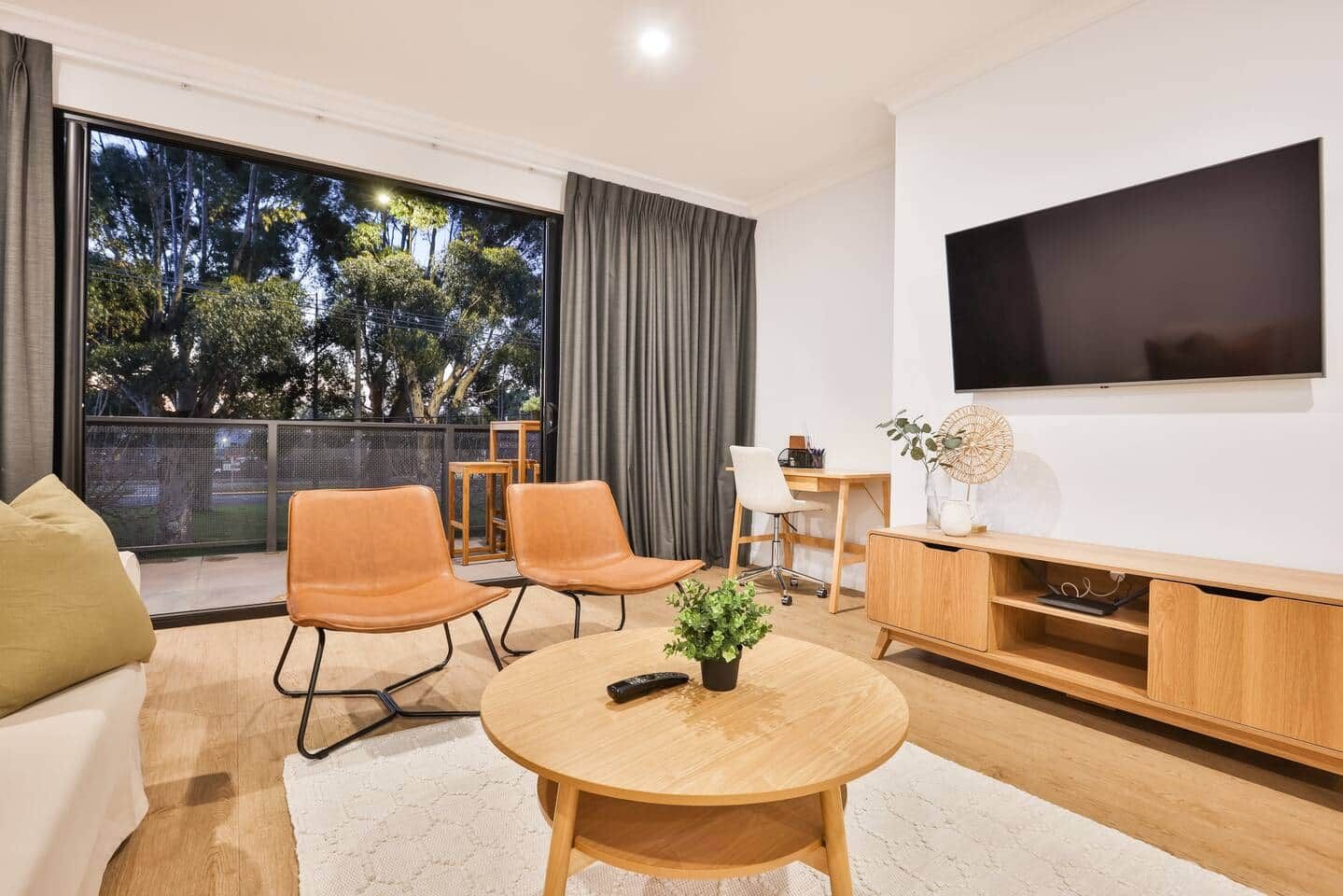 Uma sala de estar moderna com uma televisão grande, assentos confortáveis, um espaço de jantar e uma varanda com vista para árvores.