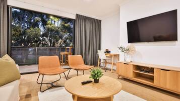 Uma sala de estar moderna com uma televisão grande, assentos confortáveis, um espaço de jantar e uma varanda com vista para árvores.