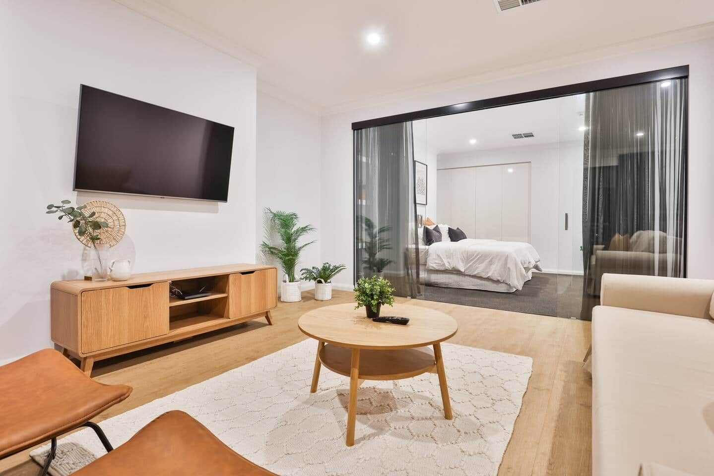 A modern living room with a flat-screen TV, wooden entertainment unit, round coffee table, and a view into the bedroom through a glass door.