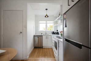 A compact kitchen with white cabinetry, a dishwasher, a stove, and a refrigerator. - Marigold by Homello (Auckland)