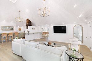 A spacious living room with a white sofa, a dining area with a brick wall, and a large flat-screen TV mounted on the wall.