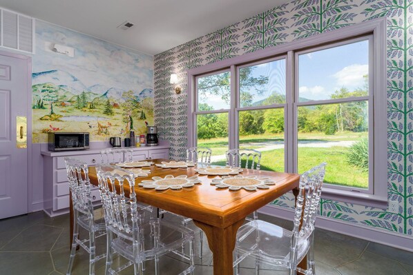 Un comedor con una mesa de madera y sillas transparentes, una cocina con microondas y cafetera, y un ventanal con vistas a un paisaje verde.