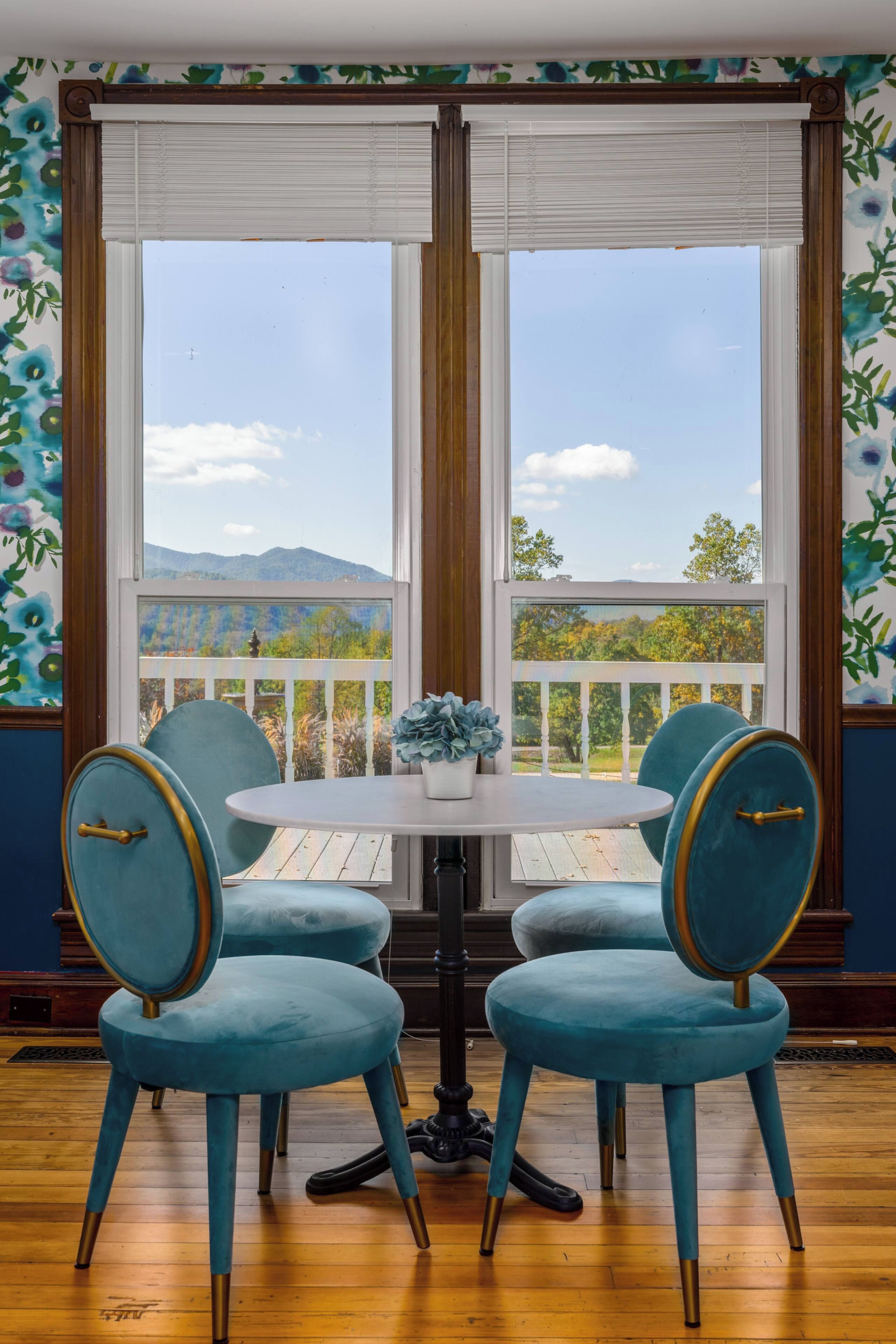 Una habitación con una mesa redonda y cuatro sillas, ventanas amplias con vistas a una montaña y árboles, y una pared decorativa con motivos florales.
