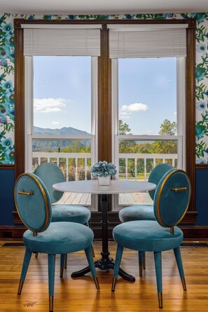 A room with a round table and four chairs, large windows with a view of a mountain and trees, and a decorative wall with a floral pattern.