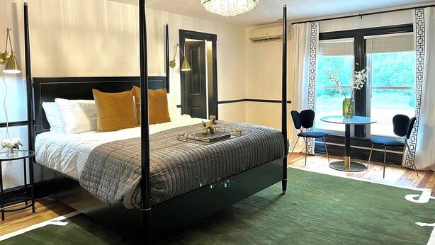 A four-poster bed with a modern design, a chandelier, and a view of the outdoors through a large window. - Everett Estate | King Suite + Twin Daybed (Nellysford)