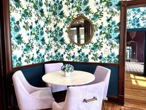 Quarto com papel de parede floral, mesa redonda com cadeiras e espelho na parede.