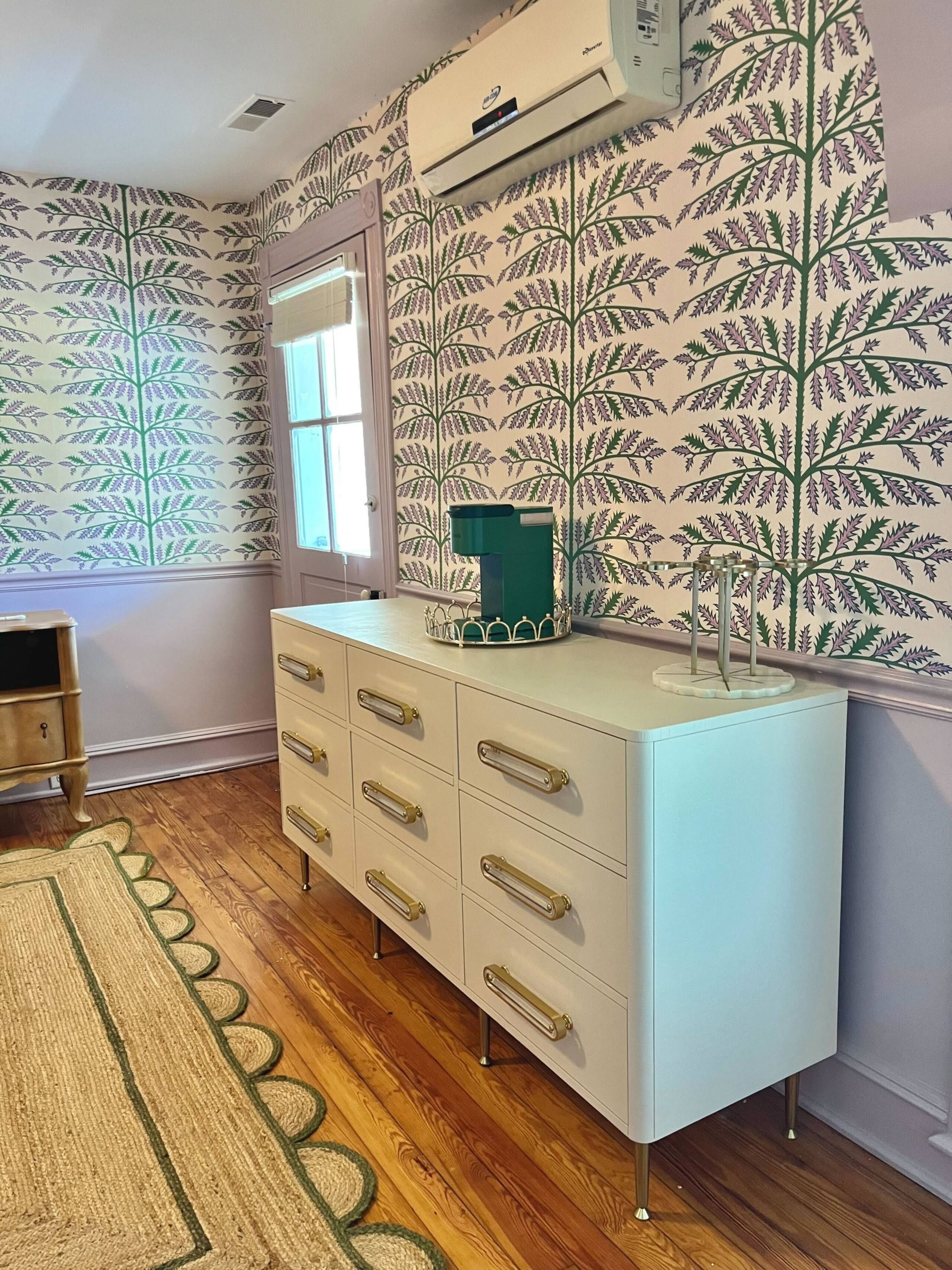 A room with a white dresser, patterned wallpaper, a wooden floor, and an air conditioning unit.