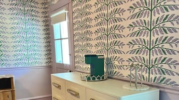 A room with a white dresser, patterned wallpaper, a wooden floor, and an air conditioning unit.