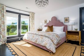 A bedroom with a large bed, a chandelier, a nightstand, and a view of a garden through the window. - Everett Estate | Mountain View King Suite (Nellysford)