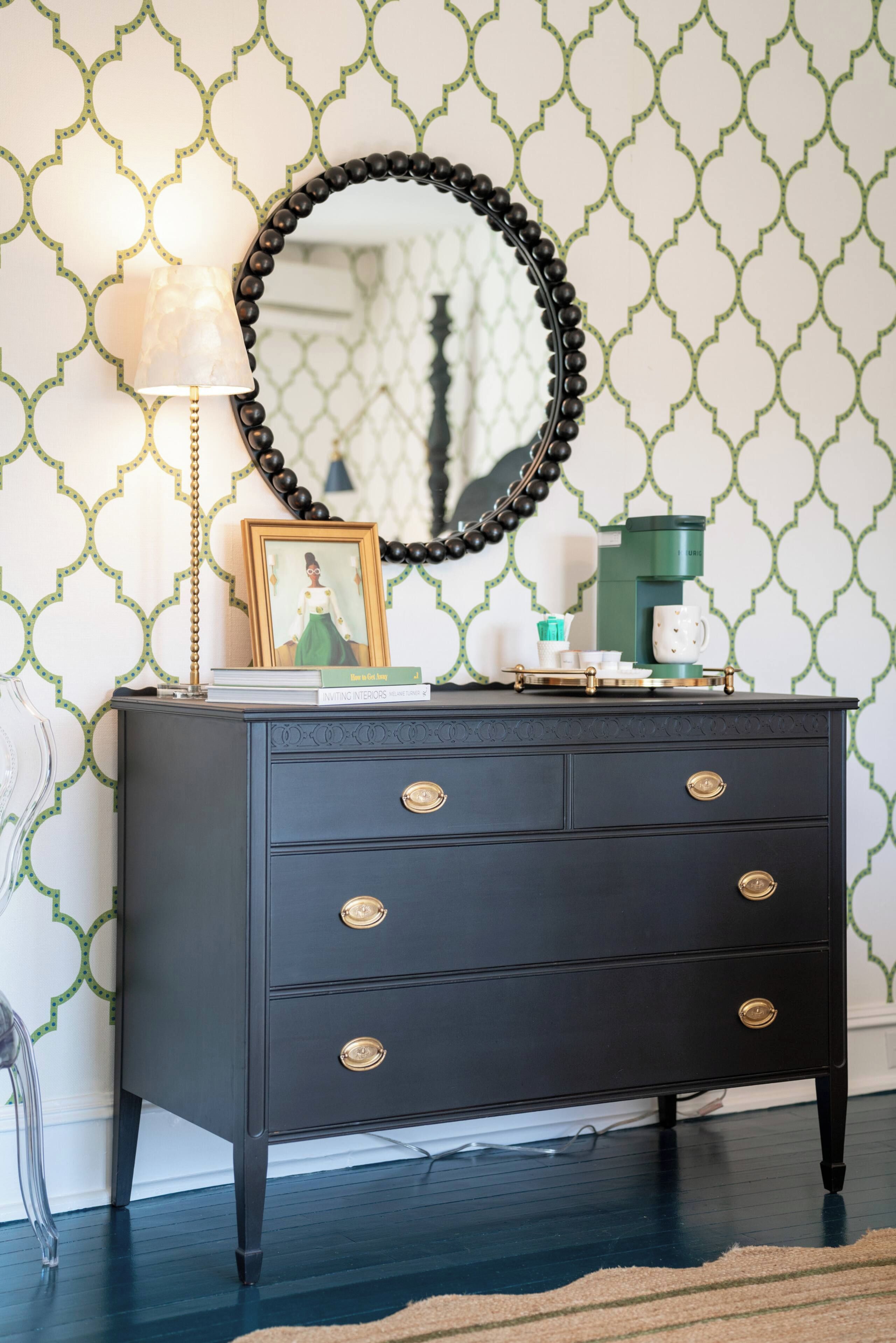 A dark wooden dresser with gold handles, a round mirror, a framed picture, and a lamp against a patterned wallpaper.
