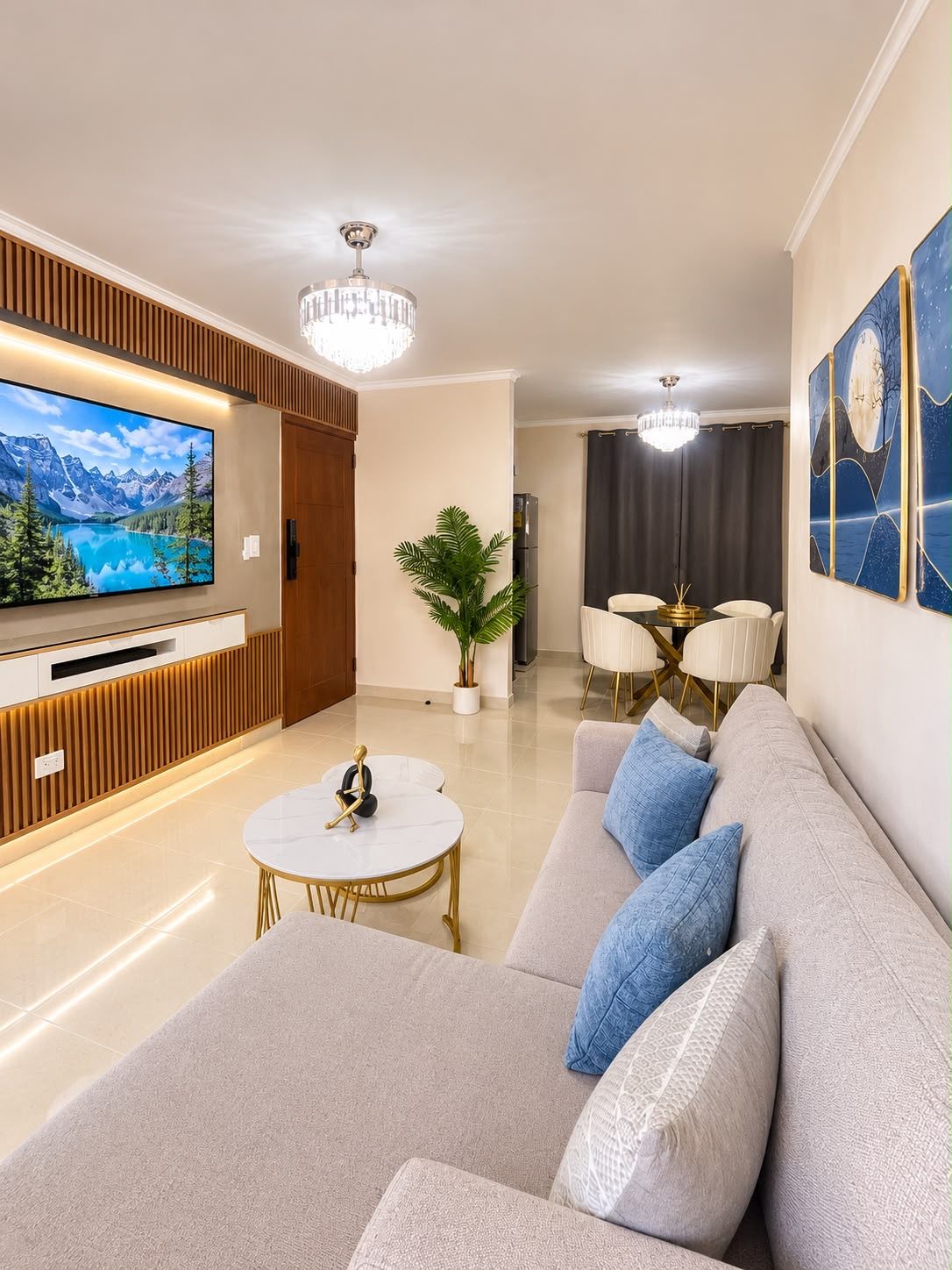A modern living room with a large flat-screen TV, a sofa, a coffee table, and a dining area with a chandelier.