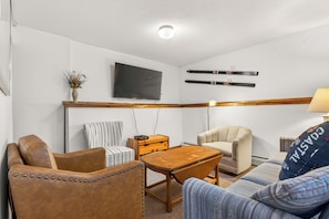 Et værelse med et fjernsyn ophængt på væggen, et træbord, to stole, en sofa og en lampe.