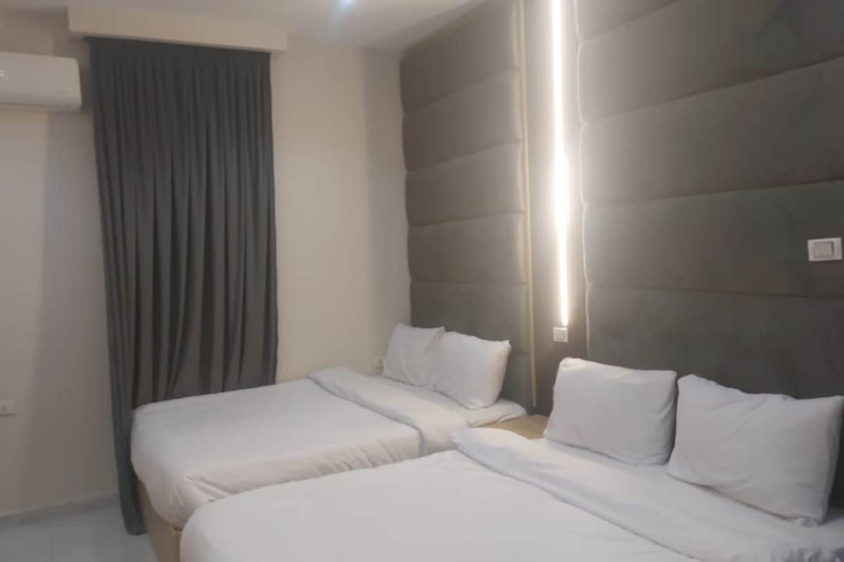 Quarto de hotel com duas camas de solteiro, uma cabeceira grande e acolchoada, uma janela com cortinas e um ar condicionado fixado na parede.
