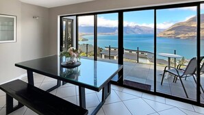 Quarto moderno com mesa de vidro e banqueta, vista para um lago e montanhas, e varanda com cadeiras.