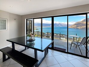 Et moderne værelse med et glasbord og bænk, udsigt til en sø og bjerge, samt en balkon med stole.