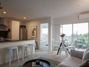 Una sala de estar moderna con un sofá, una mesa de centro y un comedor con sillas y una mesa. Cuenta con una zona de cocina con refrigerador, microondas y pileta. La habitación tiene ventanas grandes y un equipo de aire acondicionado.