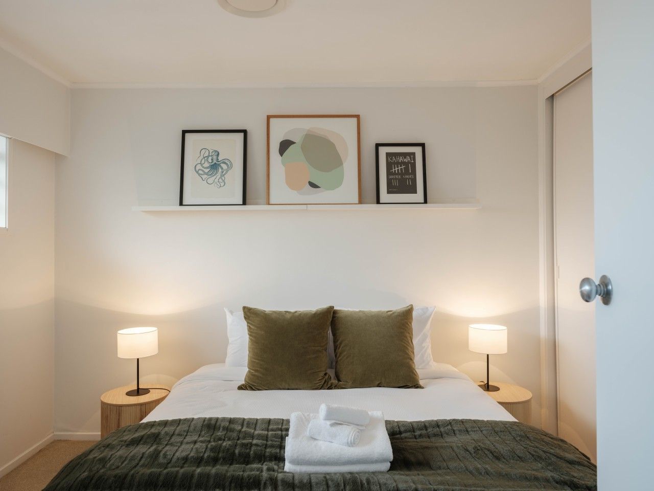 Una cama bien tendida con dos almohadas, una manta y una toalla doblada. Hay dos lámparas de noche, un estante empotrado en la pared con tres obras de arte enmarcadas y una puerta a la derecha.