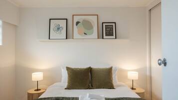 Una cama bien tendida con dos almohadas, una manta y una toalla doblada. Hay dos lámparas de noche, un estante empotrado en la pared con tres obras de arte enmarcadas y una puerta a la derecha.