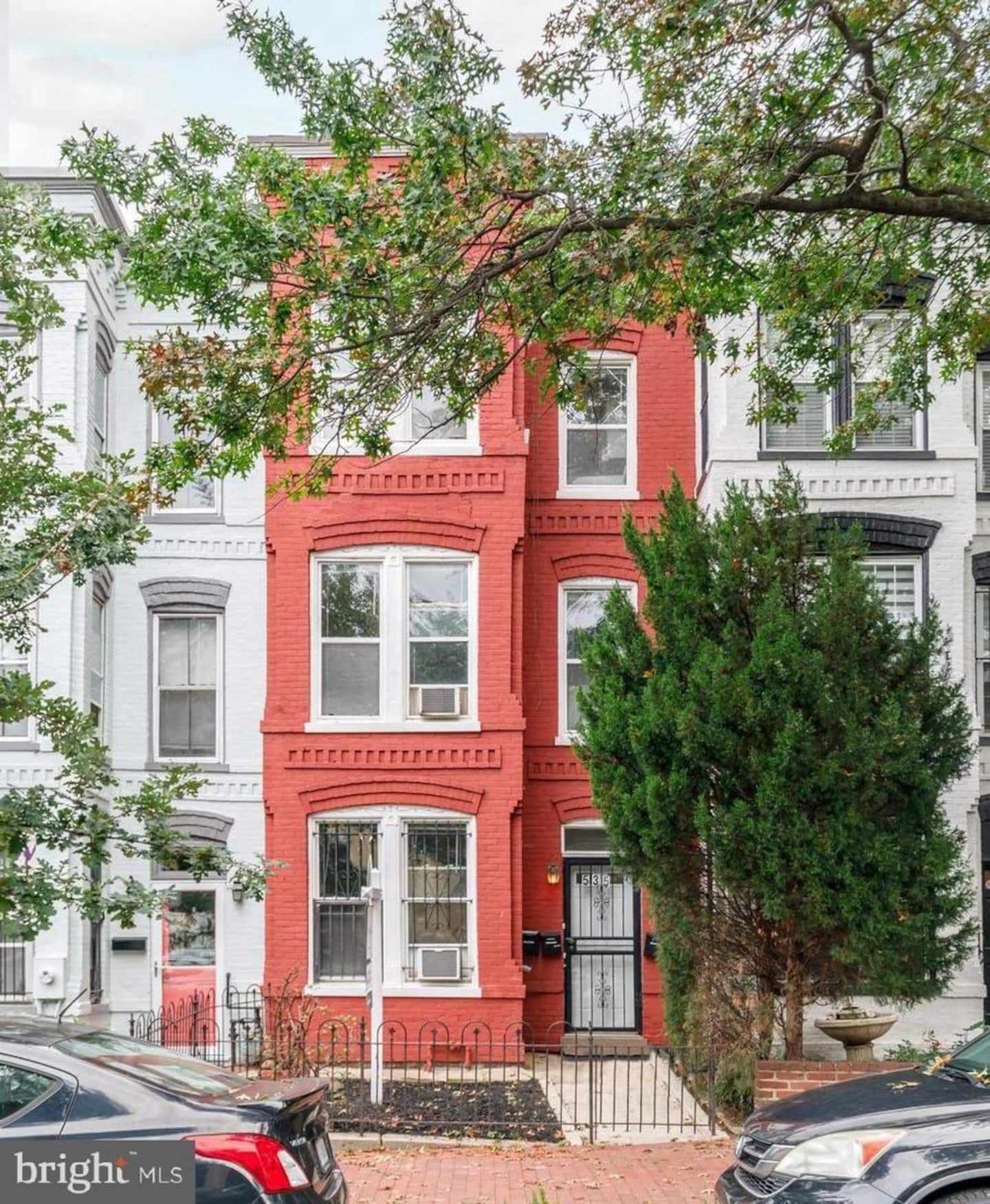 A red brick row house with white trim, a black metal fence, and a tree in the foreground.