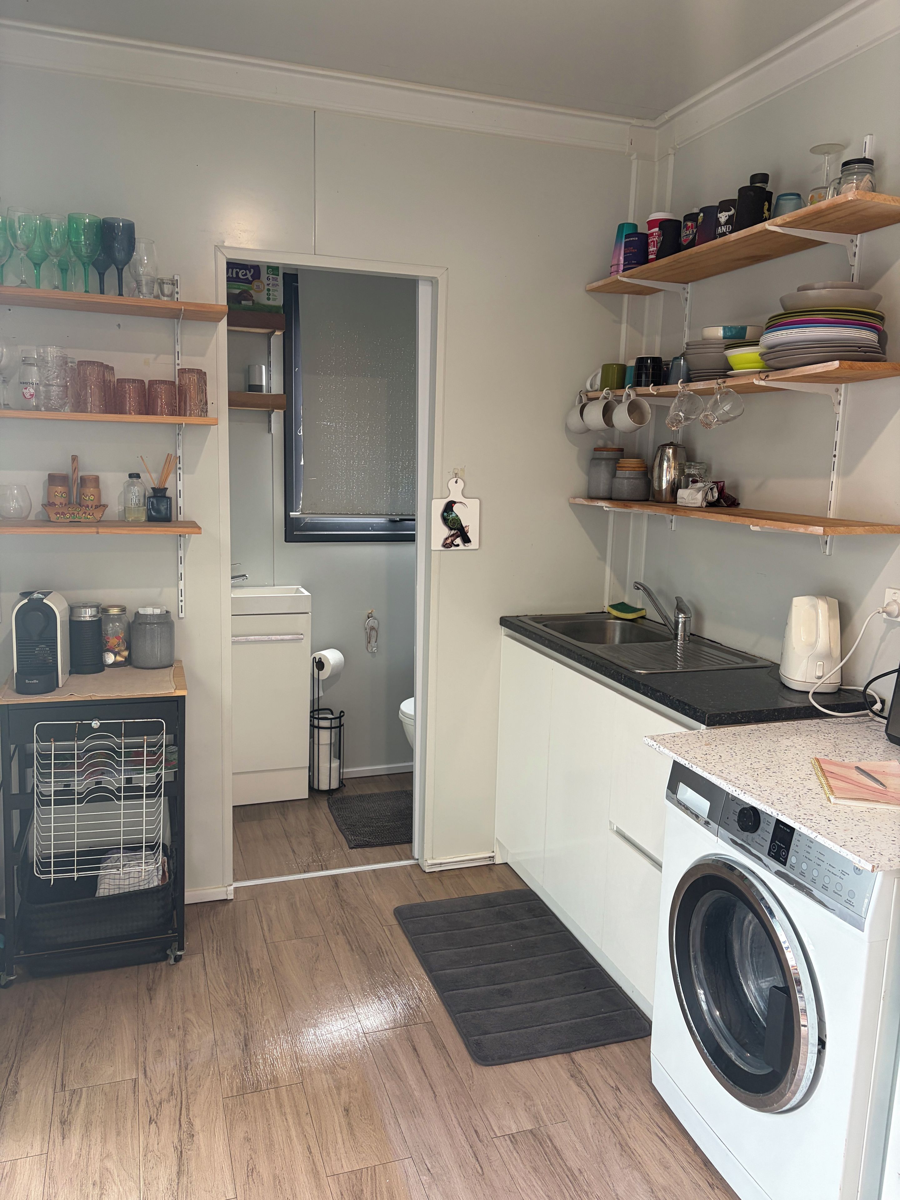 A compact kitchen with a washing machine, shelves with dishes, and a door leading to a bathroom.
