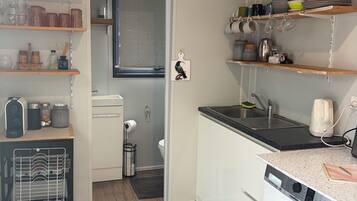 A compact kitchen with a washing machine, shelves with dishes, and a door leading to a bathroom.