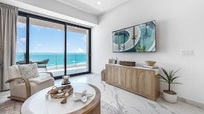 A modern living room with a large window overlooking the ocean, a round coffee table, a beige sofa, and a wooden cabinet. - Modern Oceanfront 1BR with Stunning Sea of Cortez Views (Puerto Peñasco)