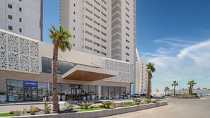 A modern hotel with a glass facade, palm trees, and a Blue Harbor sign. - Modern Oceanfront 1BR with Stunning Sea of Cortez Views (Puerto Peñasco)