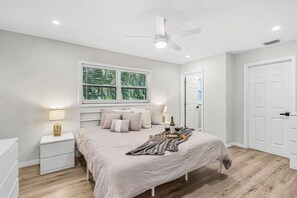 A bedroom with a bed, bedside tables, a ceiling fan, a window with blinds, and a door. - Heated Pool, Gulf Fun & Paddleboard Adventures in Cape Coral - Manatee Cottage - Roelens (Cape Coral)
