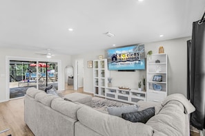 A modern living room with a large grey sectional sofa, a flat-screen TV mounted on the wall, and a white entertainment unit with decorative items. - Heated Pool, Gulf Fun & Paddleboard Adventures in Cape Coral - Manatee Cottage - Roelens (Cape Coral)